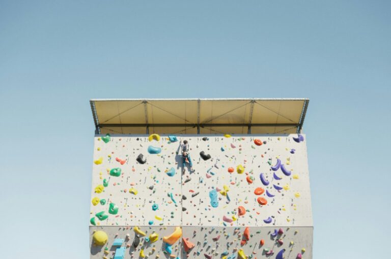 Person climbing a colorful outdoor rock wall under a clear blue sky, symbolizing willpower vs motivation and persistence in achieving goals.