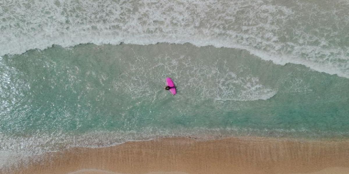 Aerial view of a person on a pink surfboard paddling in turquoise ocean waves near a sandy beach—symbolizing the mindfulness technique of urge surfing.