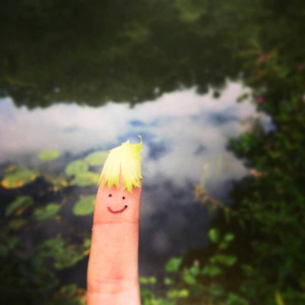 Close-up of a finger with a drawn smiling face and a small leaf as hair, held against a blurred background of water and greenery, symbolizing playfulness and the calming effect of a gentle smile.