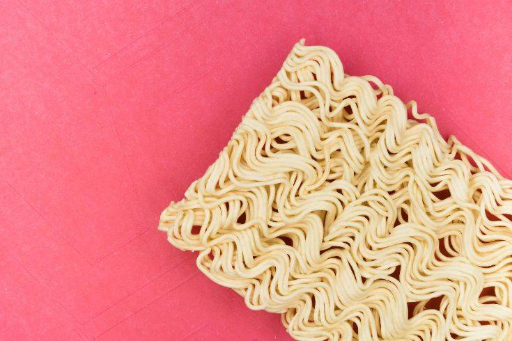 A block of uncooked instant noodles on a pink background, symbolizing the 'wet noodle technique' for stress reduction, which encourages relaxing the body like limp noodles to release tension.