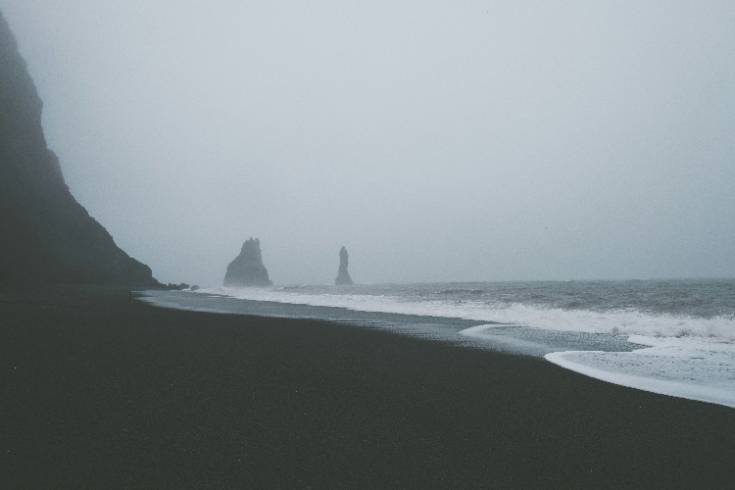 A foggy coastal landscape with dark sand, gentle waves, and distant rock formations, creating a calm, reflective atmosphere for a blog about meeting disappointment with compassion.