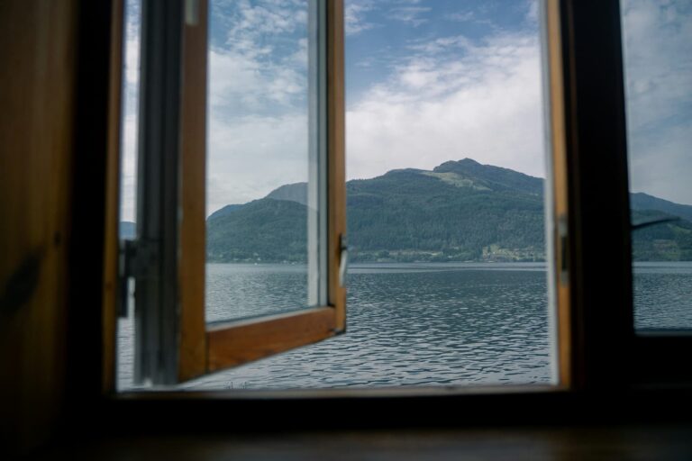 Open window overlooking a calm lake with mountains in the distance beneath a partly cloudy sky.
